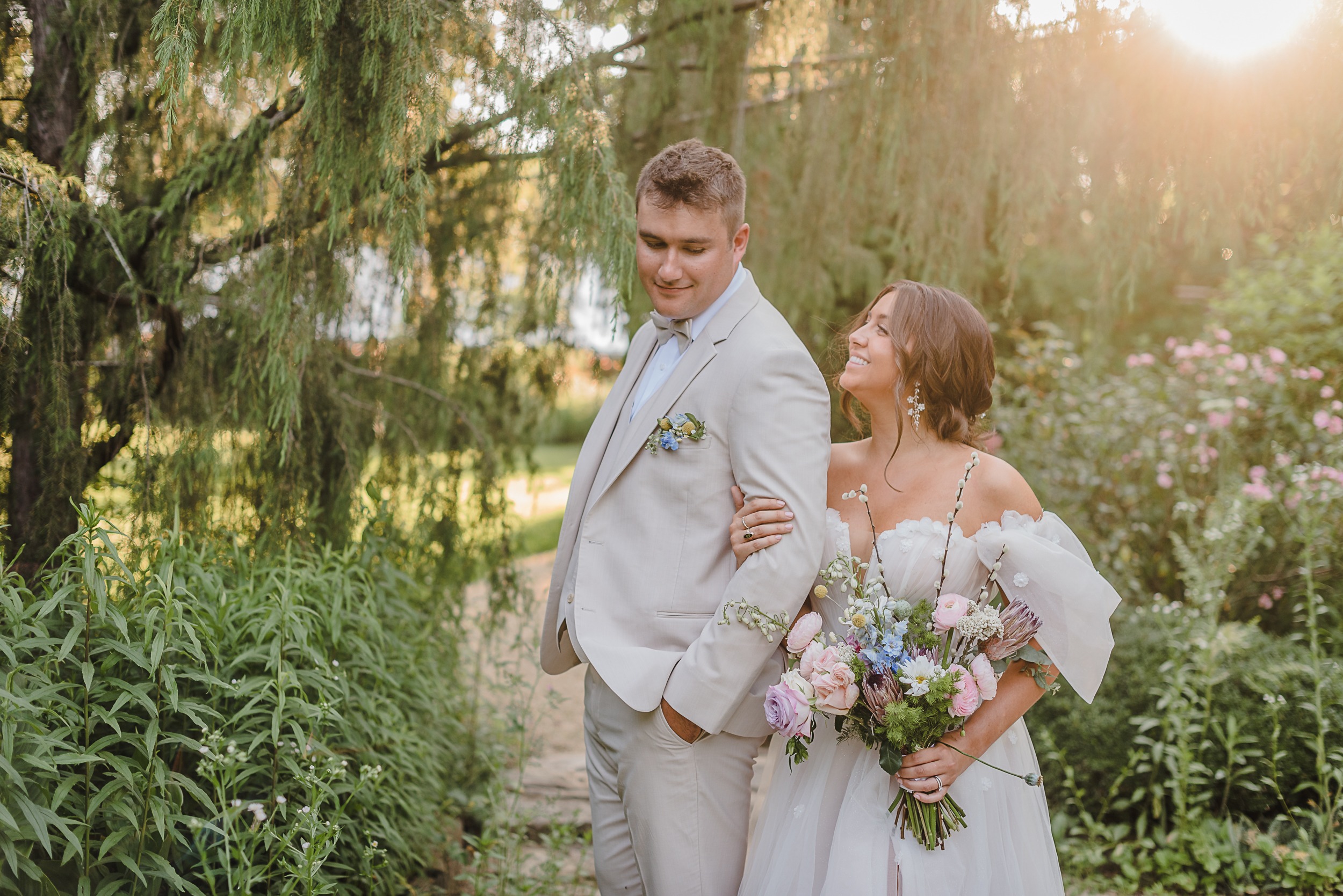 Bride looking up at Groom while holding his arm, beautiful sunlight entering behind them in a secret garden